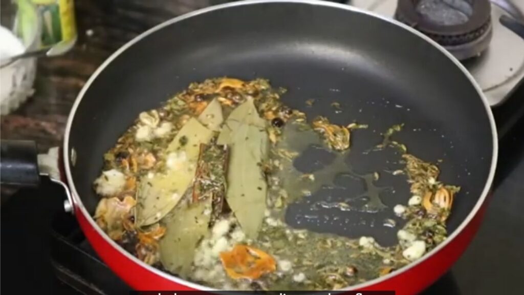 A non-stick pan with a teaspoon of olive oil, where cumin seeds, a cinnamon stick, cardamom pods, and a bay leaf sizzle, releasing aromatic smoke against a blurred kitchen stove backdrop.